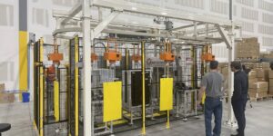 Automated panel painting and drying station inside a warehouse, with orange spray/dryer heads on a white overhead conveyor enclosed by black mesh safety fencing, and two workers observing.